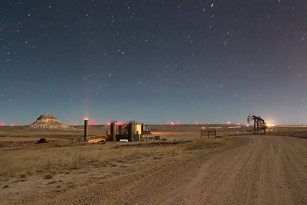 Standing on a newly cut dirt road, stars rotate over a recently constructed oil well pad with a glowing VOC combustor (volatile organic compound), heater/treaters, separators and storage tanks with one of the Pawnee Buttes in the Pawnee National Grassland in the background. Lights in the distance are from miles of wind turbines.